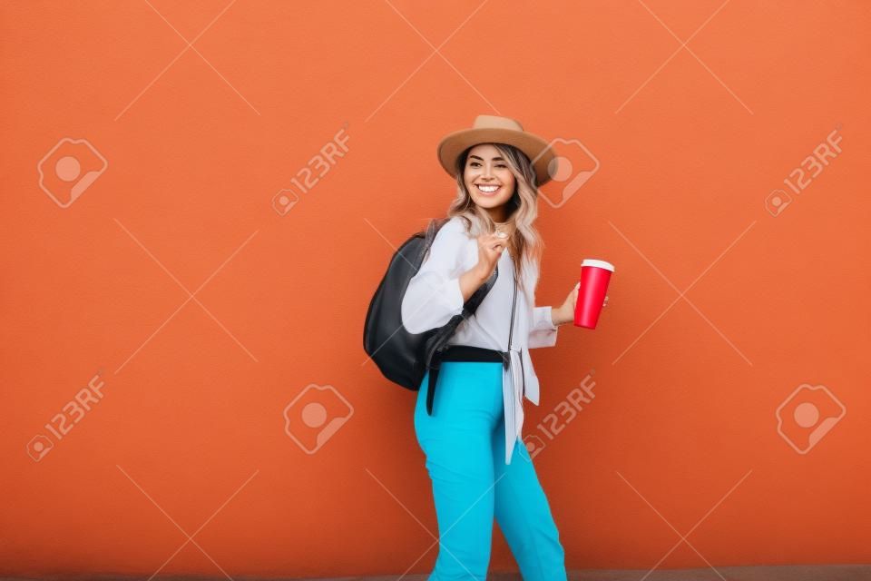 Porträt einer aktiven, stilvollen Frau in hellem Hemd mit Hut und Kaffeetasse auf dem roten Wandhintergrund im Freien. Sorgloses Lifestyle-, Kaffee- und Damenmodekonzept