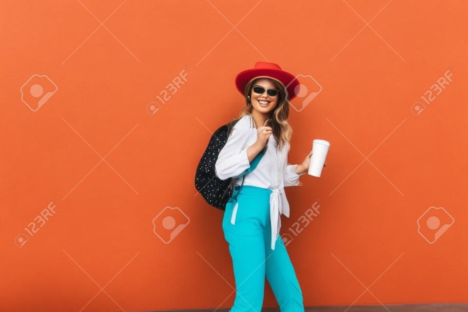 Retrato de una mujer elegante y activa vestida con un pantalones brillante con sombrero y una taza de café en el fondo de la pared roja al aire libre. Concepto de estilo de vida sin preocupaciones, café y moda femenina