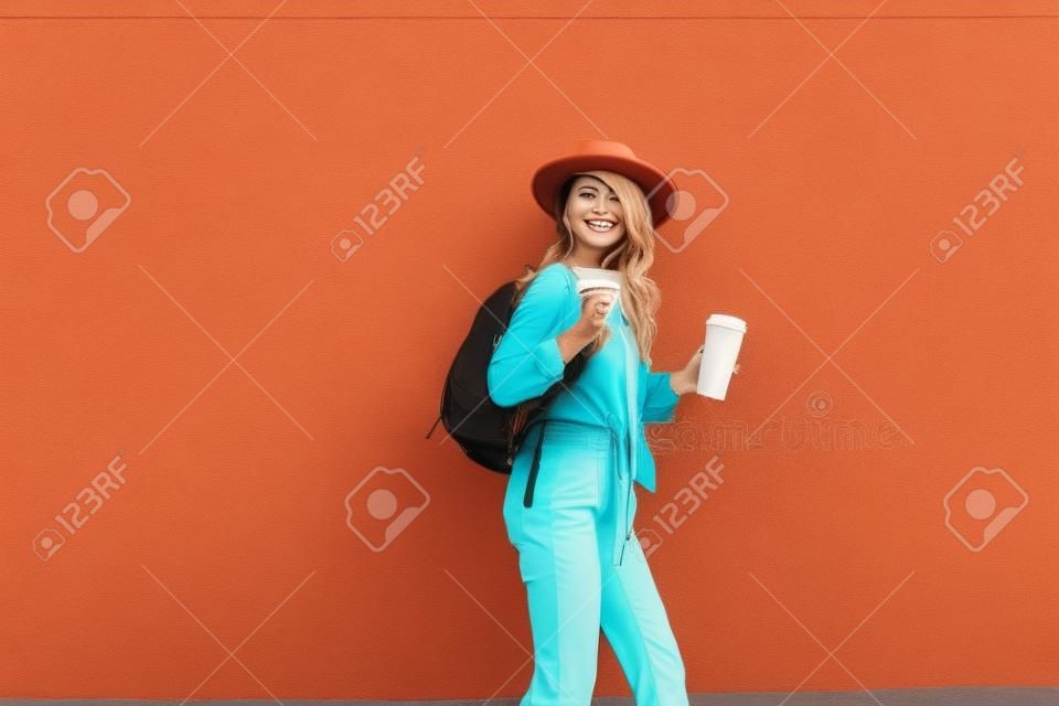 Porträt einer aktiven, stilvollen Frau in hellem Hemd mit Hut und Kaffeetasse auf dem roten Wandhintergrund im Freien. Sorgloses Lifestyle-, Kaffee- und Damenmodekonzept