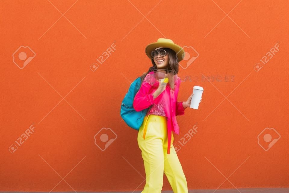 Retrato de una mujer elegante y activa vestida con un pantalones brillante con sombrero y una taza de café en el fondo de la pared roja al aire libre. Concepto de estilo de vida sin preocupaciones, café y moda femenina