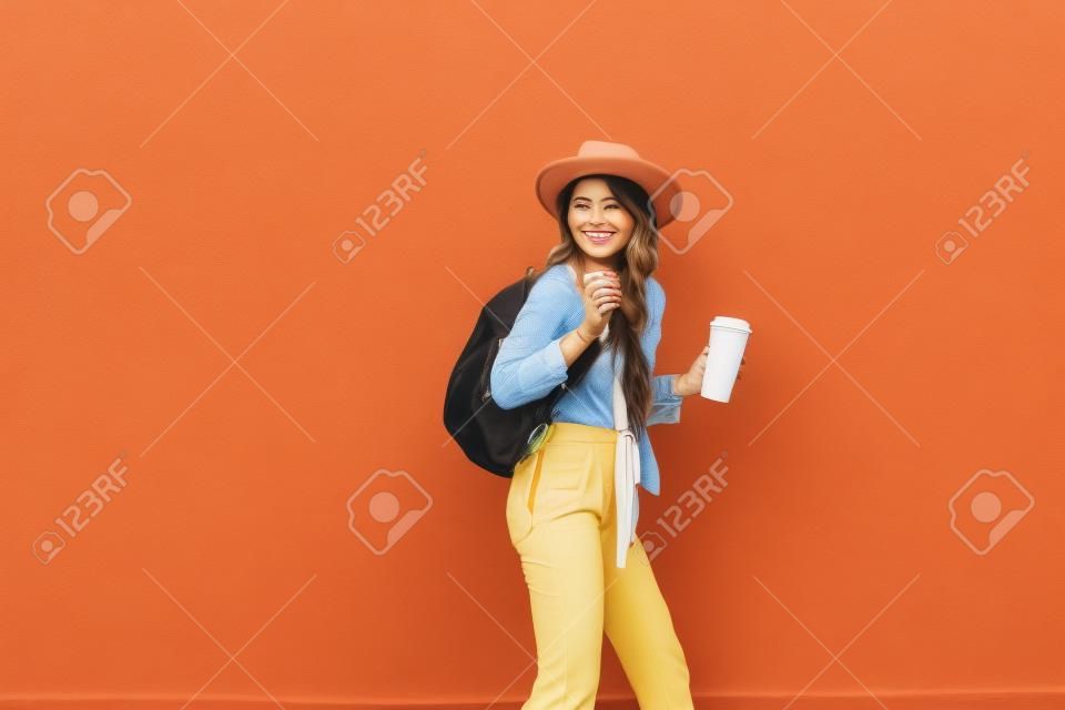 Portrait d'une femme élégante active vêtue d'une chemise lumineuse avec un chapeau et une tasse de café sur le fond du mur rouge à l'extérieur. Style de vie insouciant, café et concept de mode pour femmes