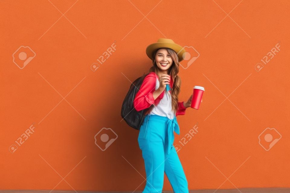 야외에서 빨간 벽 배경에 모자와 커피 컵이 달린 밝은 셔츠를 입은 활동적인 세련된 여성의 초상화. 평온한 라이프 스타일, 커피 및 여성 패션 컨셉