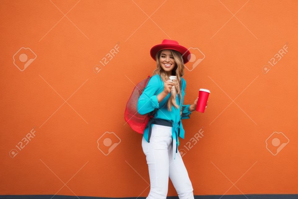 Portrait d'une femme élégante active vêtue d'une chemise lumineuse avec un chapeau et une tasse de café sur le fond du mur rouge à l'extérieur. Style de vie insouciant, café et concept de mode pour femmes