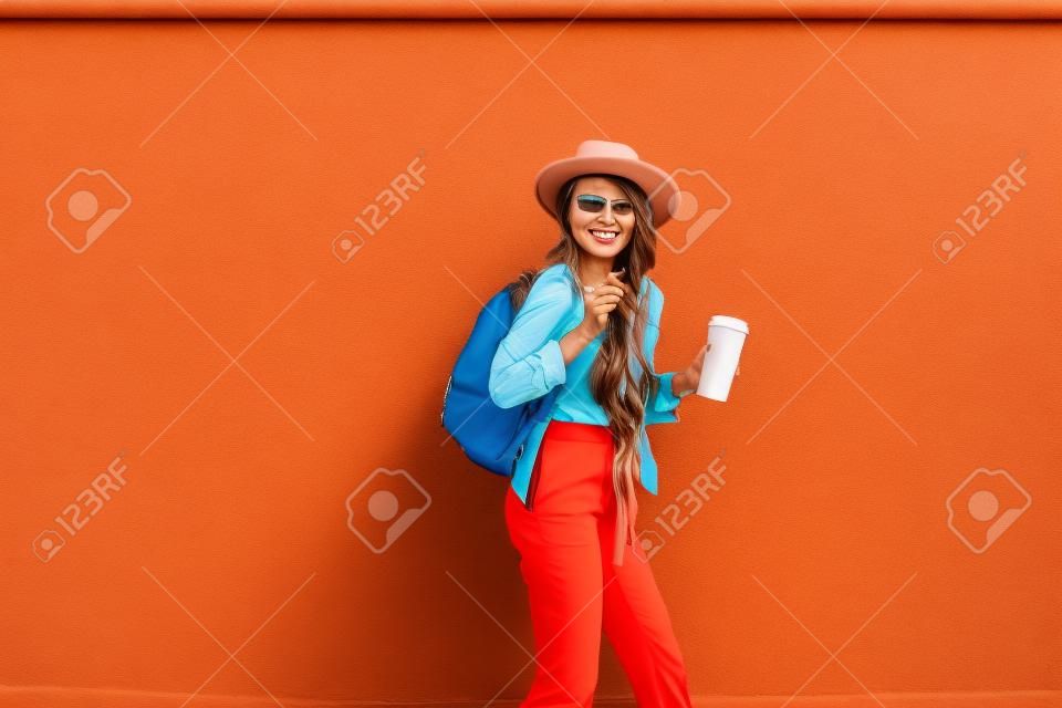 Ritratto di una donna alla moda attiva vestita con una camicia luminosa con cappello e tazza di caffè sullo sfondo rosso della parete all'aperto. Stile di vita spensierato, caffè e concetto di moda femminile