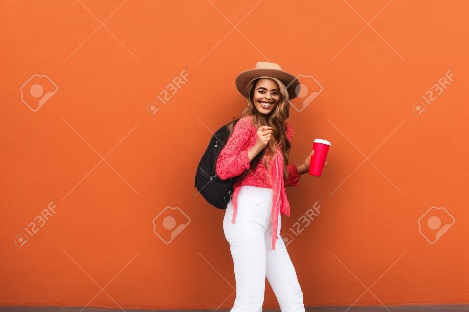 야외에서 빨간 벽 배경에 모자와 커피 컵이 달린 밝은 셔츠를 입은 활동적인 세련된 여성의 초상화. 평온한 라이프 스타일, 커피 및 여성 패션 컨셉