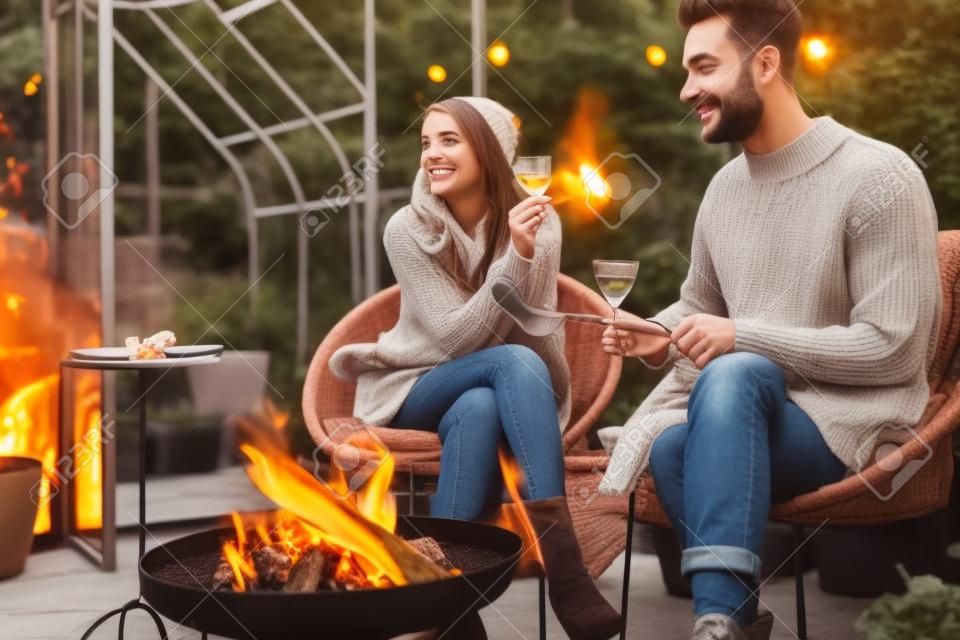 Pareja joven y elegante asando comida y calentándose mientras se sientan juntos junto al fuego, pasando la noche de otoño en un ambiente acogedor en el jardín