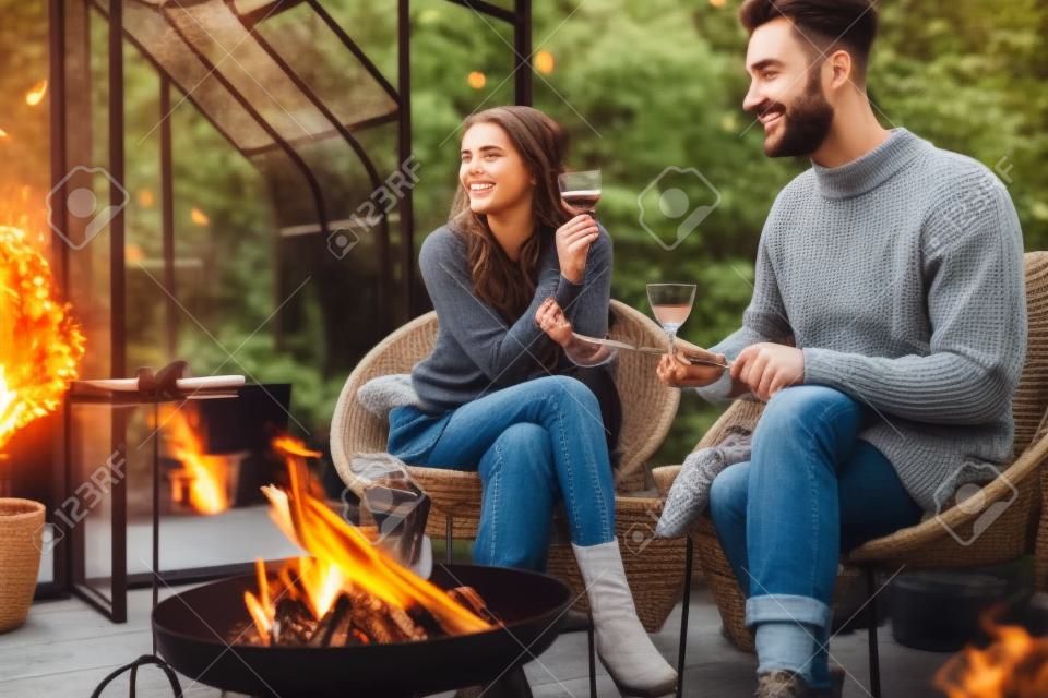 Jeune couple élégant grillant de la nourriture et s'échauffant tout en étant assis ensemble près du feu, passant une soirée d'automne dans une atmosphère chaleureuse dans le jardin