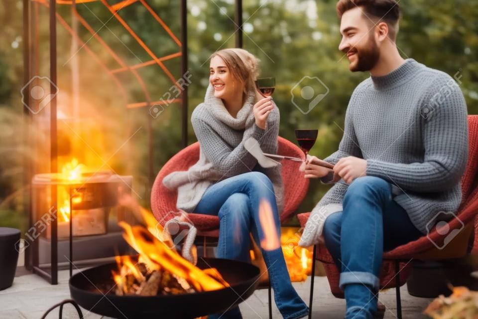 Junges, stilvolles Paar grillt Essen und wärmt sich auf, während es gemeinsam am Feuer sitzt und den Herbstabend in gemütlicher Atmosphäre im Garten verbringt