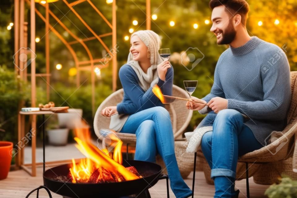 火のそばに座って食べ物を焼き、ウォーミングアップし、庭の居心地の良い雰囲気の中で秋の夜を過ごす若いスタイリッシュな夫婦