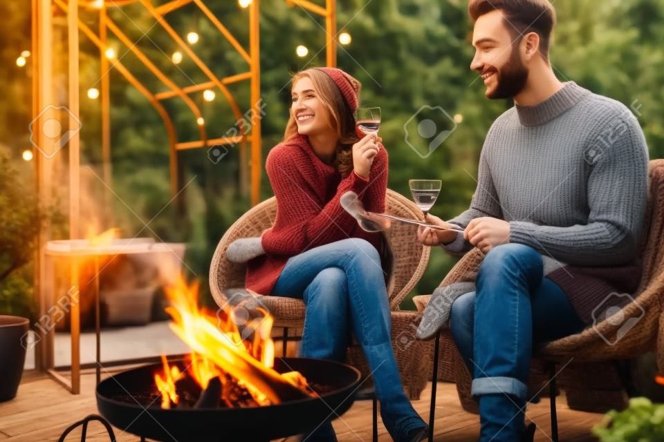 Giovane coppia elegante che griglia cibo e si riscalda mentre si siede insieme accanto al fuoco, trascorrendo le serate autunnali in un'atmosfera accogliente in giardino