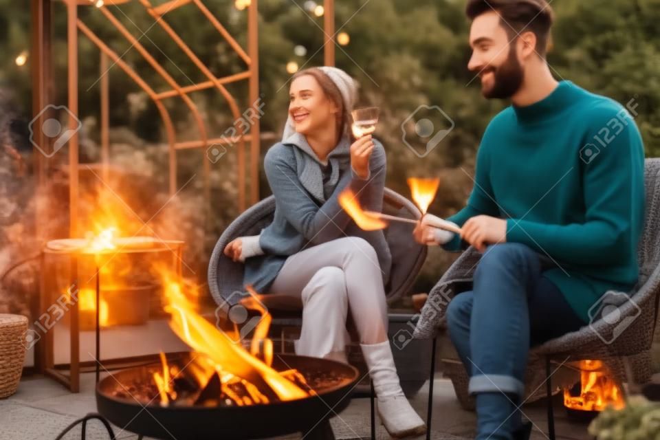 Jovem casal elegante grelhando comida e aquecendo enquanto sentado junto ao fogo, passando o outono à noite no ambiente acolhedor no jardim