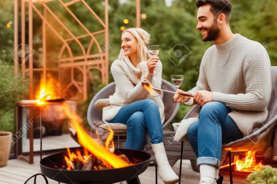 Junges, stilvolles Paar grillt Essen und wärmt sich auf, während es gemeinsam am Feuer sitzt und den Herbstabend in gemütlicher Atmosphäre im Garten verbringt