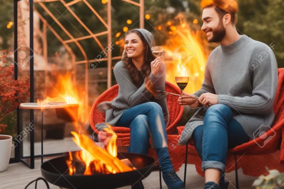 Jovem casal elegante grelhando comida e aquecendo enquanto sentado junto ao fogo, passando o outono à noite no ambiente acolhedor no jardim