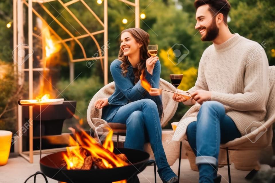 Giovane coppia elegante che griglia cibo e si riscalda mentre si siede insieme accanto al fuoco, trascorrendo le serate autunnali in un'atmosfera accogliente in giardino