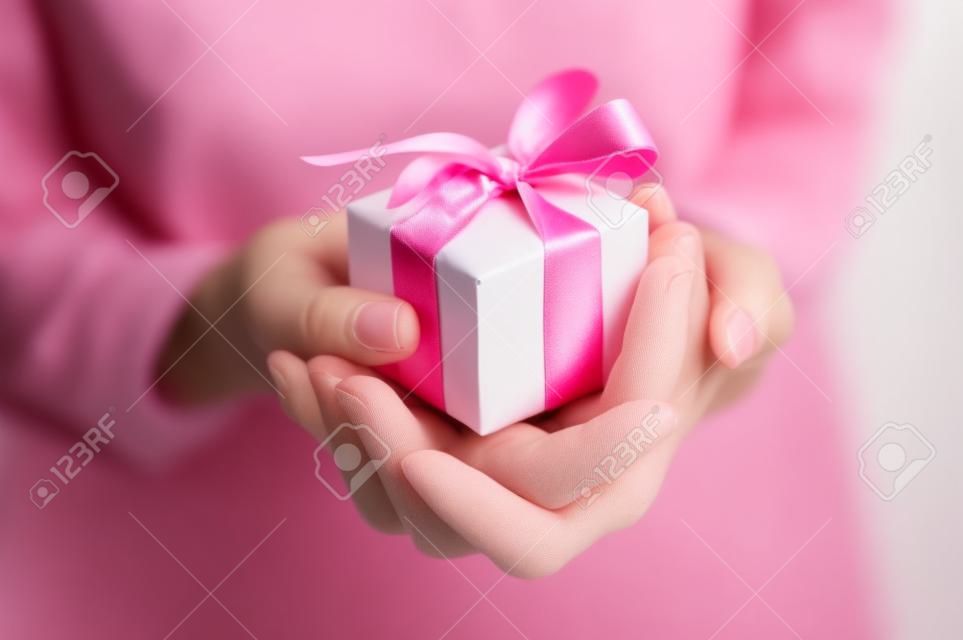 Close up tiro de mãos femininas segurando um pequeno presente envolto com fita rosa. Pequeno presente nas mãos de uma mulher dentro. Profundidade rasa do campo com foco na pequena caixa.