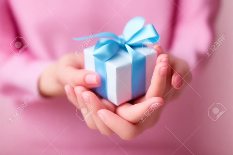 Закрыть выстрел из женских руках небольшой подарок, завернутый с розовой лентой. Малый подарок в руках женщины в помещении. Малая глубина резкости с акцентом на маленькой коробке.