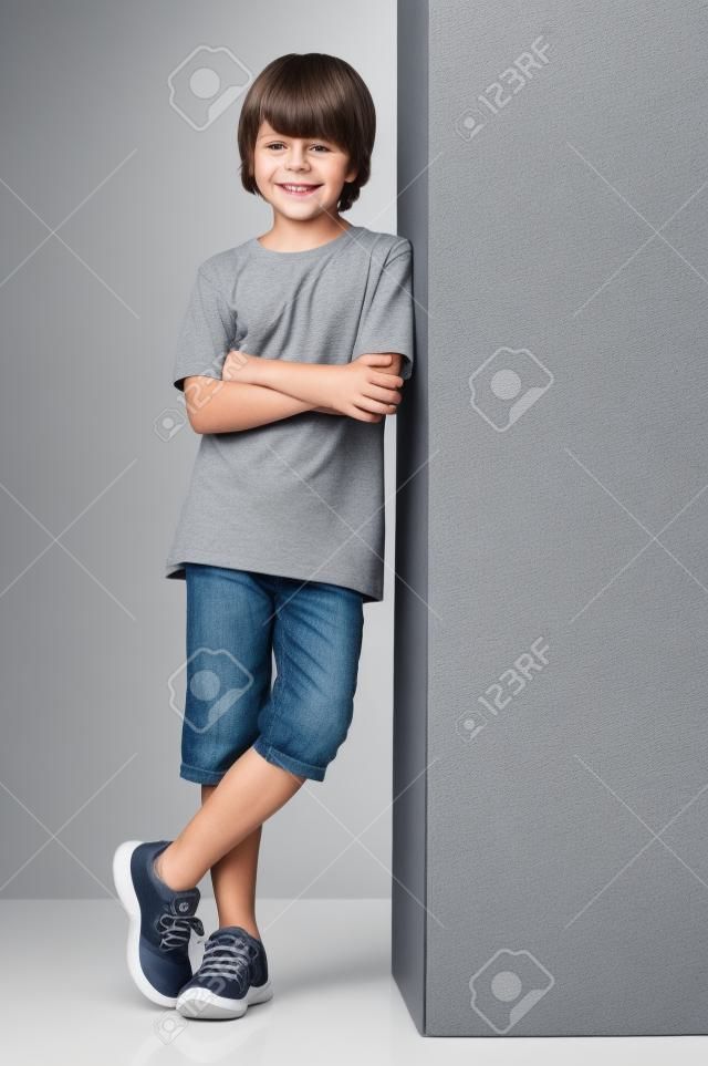 Sourire petit garçon posant contre le mur gris isolé sur fond blanc. Heureux enfant mignon debout contre un fond blanc. Jeune garçon appuyé contre un signe gris et regardant la caméra avec les bras croisés.