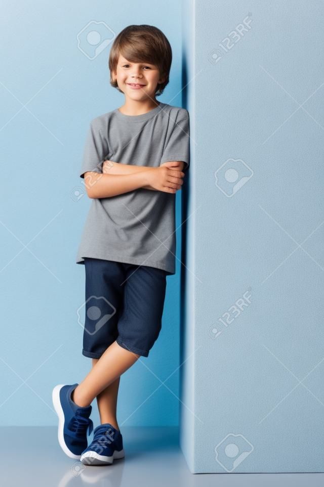 微笑的小男孩對冒充隔絕在白色背景灰色的牆。快樂可愛的孩子站在白色背景。小男孩靠在灰色的跡象，看著攝像頭，雙臂交叉。