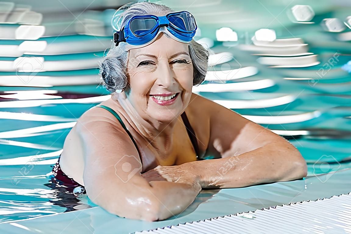 成熟女子戴游泳眼镜在游泳池适合活跃的高级女子享受退休站在游泳池和看相机高兴的高级健康老太太享受积极的生活方式