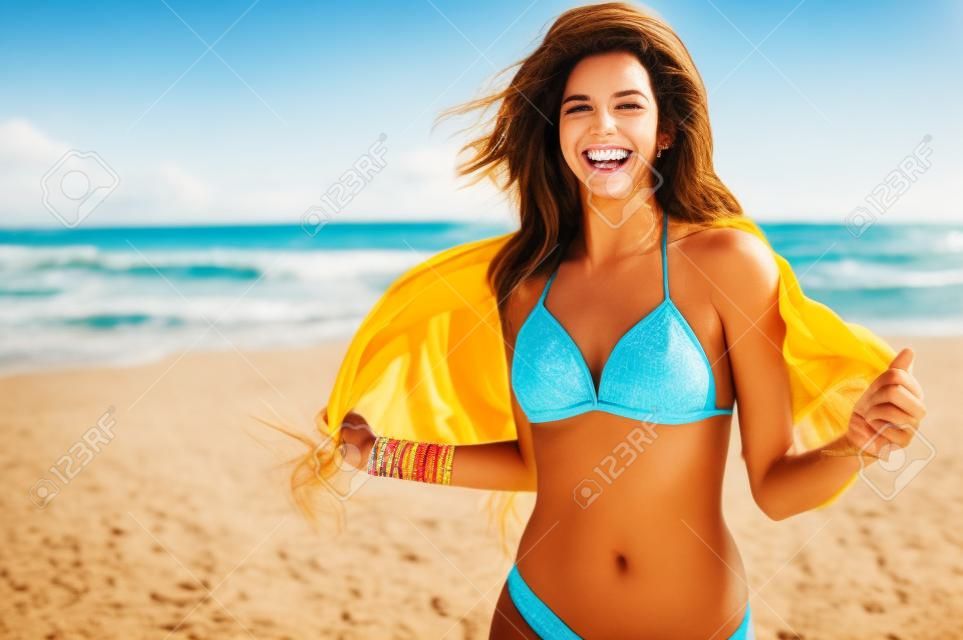 Glückliche Frau, die Spaß am Strand lächelt und hat. Sommerporträt des jungen schönen Mädchens, das auf Strand mit einem gelben Schal läuft. Lateinisches Mädchen, das Kamera mit Freude lacht und betrachtet.