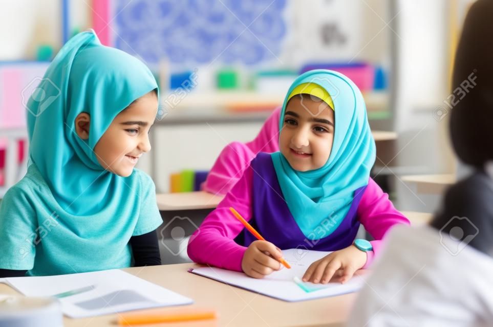 Young arab girl with hijab doing exercise with her bestfriend at international school. Asian muslim school girl sitting near her classmate during lesson. Multiethnic elementary students in classroom.  