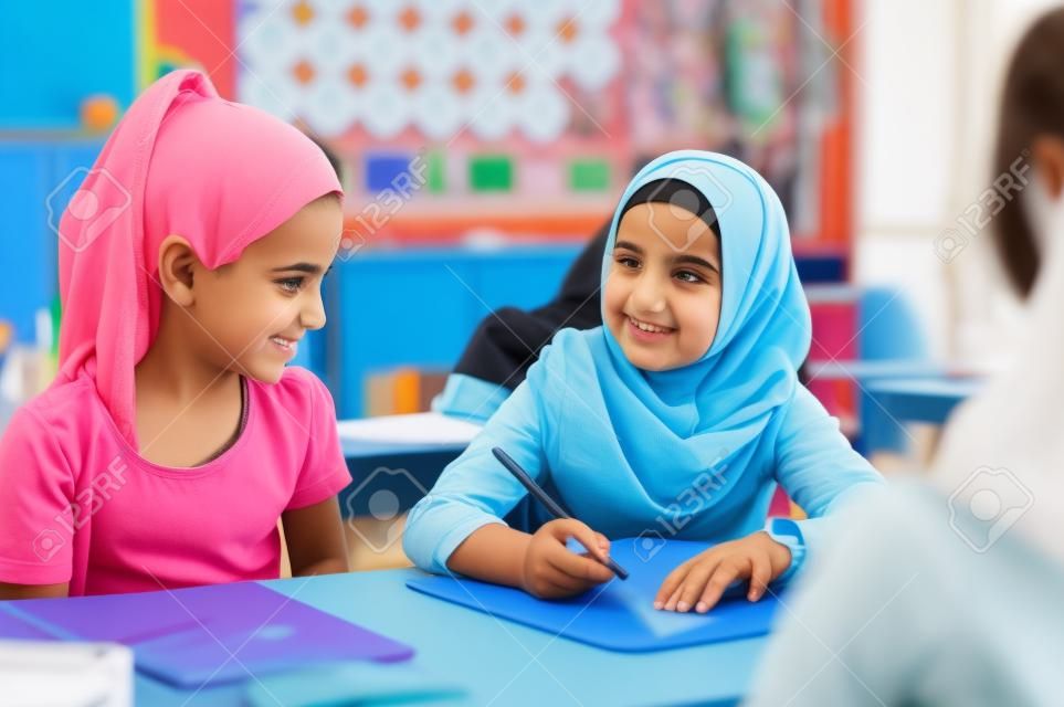 Young arab girl with hijab doing exercise with her bestfriend at international school. Asian muslim school girl sitting near her classmate during lesson. Multiethnic elementary students in classroom.  