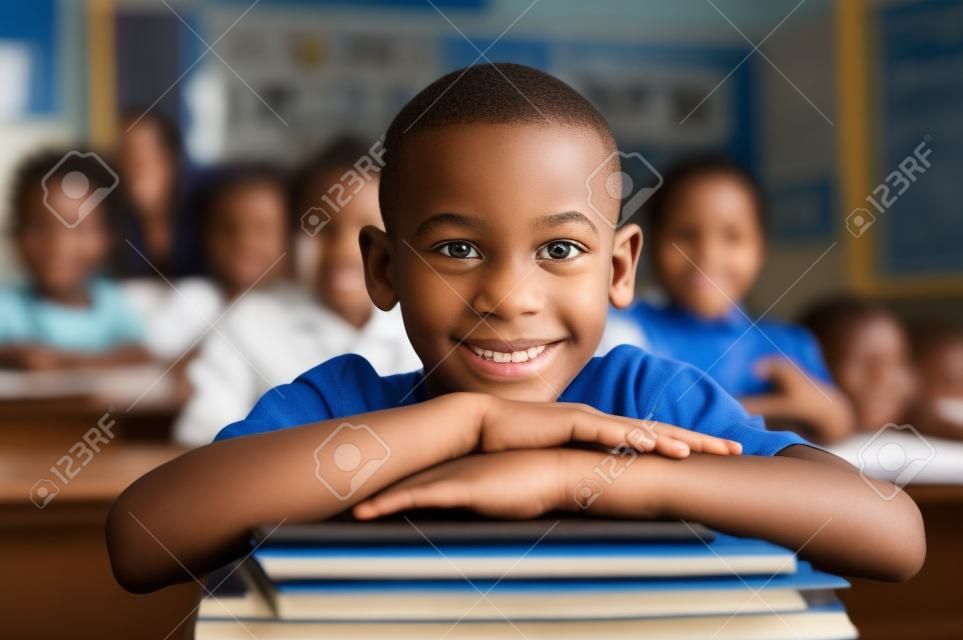 Portret van Afrikaanse Amerikaanse schooljongen leunend op bureau met klasgenoten op de achtergrond. Gelukkig jong kind zitten en leunende kin op gestapelde boeken in de klas. Portret van elementaire leerling kijkend naar camera.