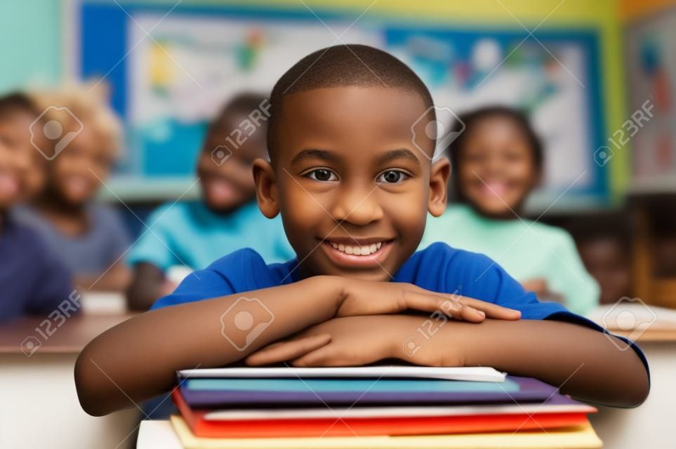 倾斜在有同学的书桌的非裔美国人的男小学生画象在背景中。愉快的小孩和倾斜的下巴坐被堆积的书在教室。看照相机的基本的学生画象。