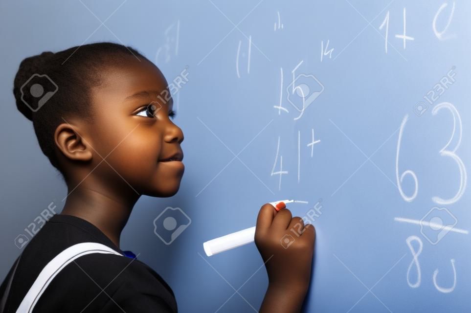 Retrato de niña africana escribiendo solución de sumas en la pizarra en la escuela. Colegiala negra resolviendo suma de suma en pizarra blanca con rotulador. Niño de la escuela pensando mientras hace problemas de matemáticas.