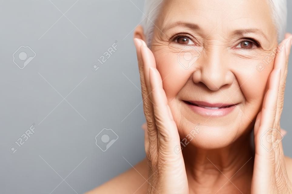 Portret van mooie senior vrouw aanraken haar perfecte huid en kijken naar camera. Close-up gezicht van volwassen vrouw met rimpels masseren gezicht geïsoleerd over grijze achtergrond. Veroudering proces concept.