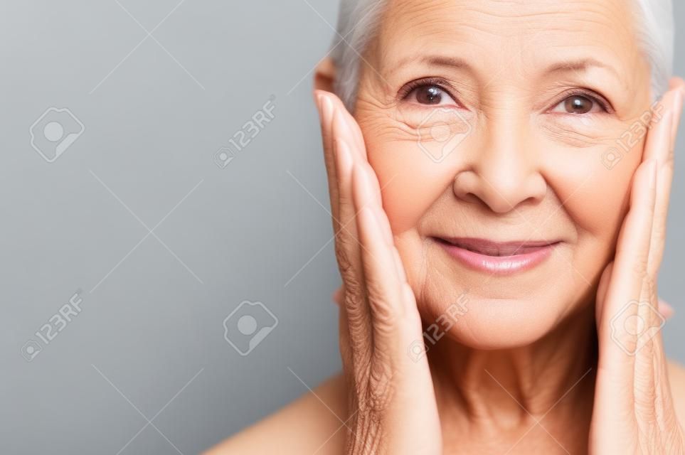 Portret pięknej starszej kobiety dotykając jej idealnej skóry i patrząc na kamery. Zbliżenie twarzy dojrzałej kobiety z zmarszczkami masowania twarzy na białym tle nad szarym tłem. Koncepcja procesu starzenia.