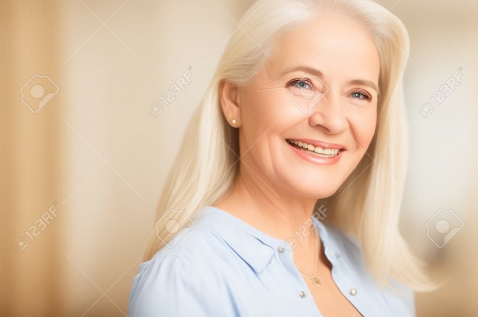 Porträt der reifen Frau, die das Leben nach dem Ruhestand genießt und Kamera betrachtet. Nahaufnahmegesicht der glücklichen älteren Frau mit dem blonden Haar lächelnd. Schöne lächelnde Dame drinnen.