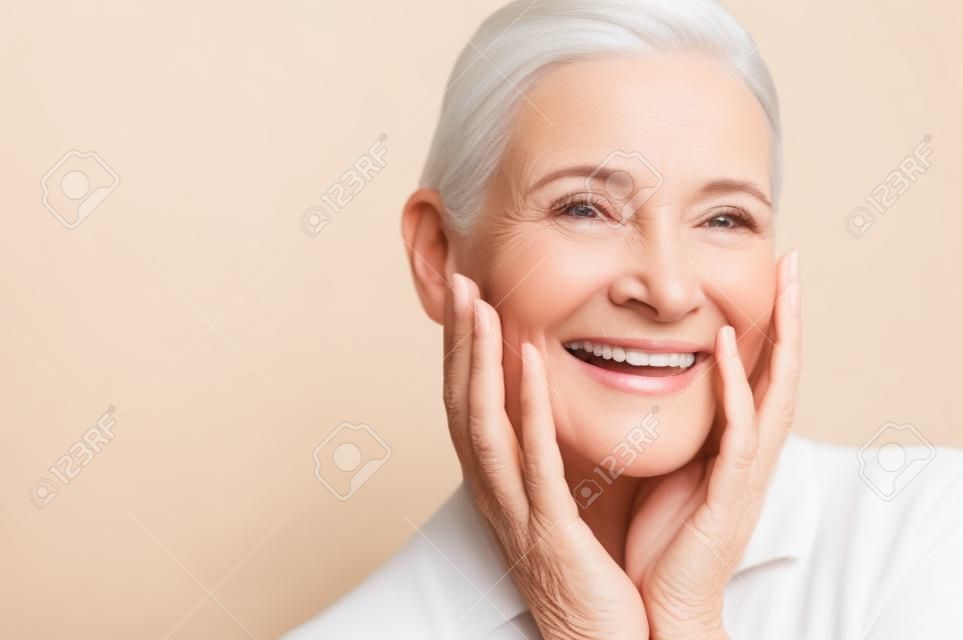 Piękno portret dojrzałej kobiety uśmiechając się ręką na twarzy. Zbliżenie twarzy szczęśliwa starsza kobieta czuje się świeżo po leczeniu przeciwstarzeniowym. Uśmiechający się piękna patrząc na kamery, dotykając jej doskonałej skóry.