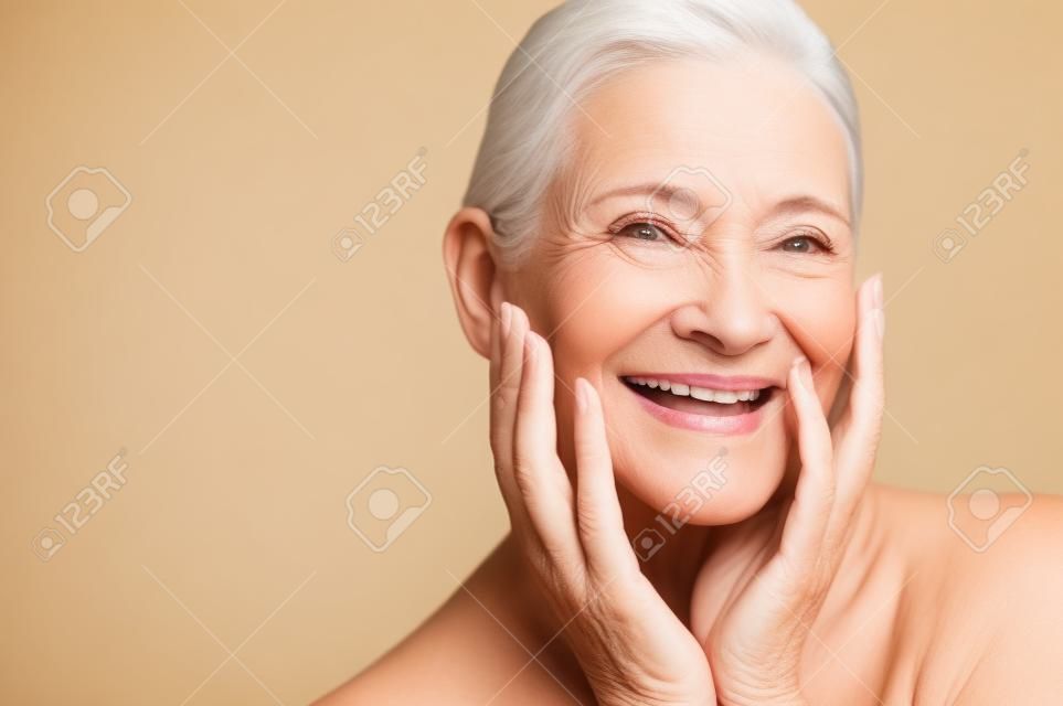微笑用在面孔的手的成熟婦女秀麗畫象。感到愉快的資深的婦女的特寫鏡頭面孔新鮮在抗衰老治療以後。看照相機的微笑的秀麗，當接觸她完善的皮膚時。
