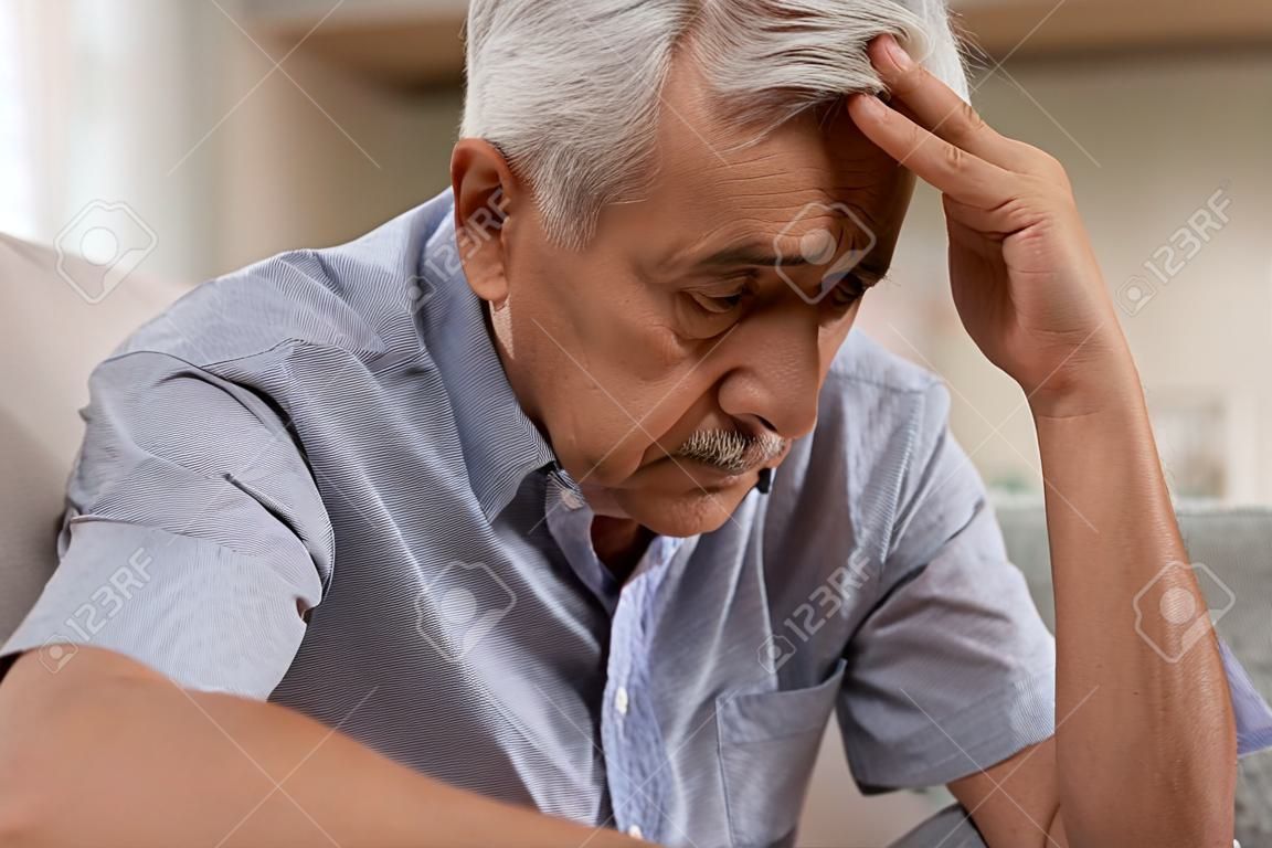 Thoughtful senior man sitting on couch. Depressed sad man sitting with hand on head thinking while looking away. Elderly man suffering from alzheimer.