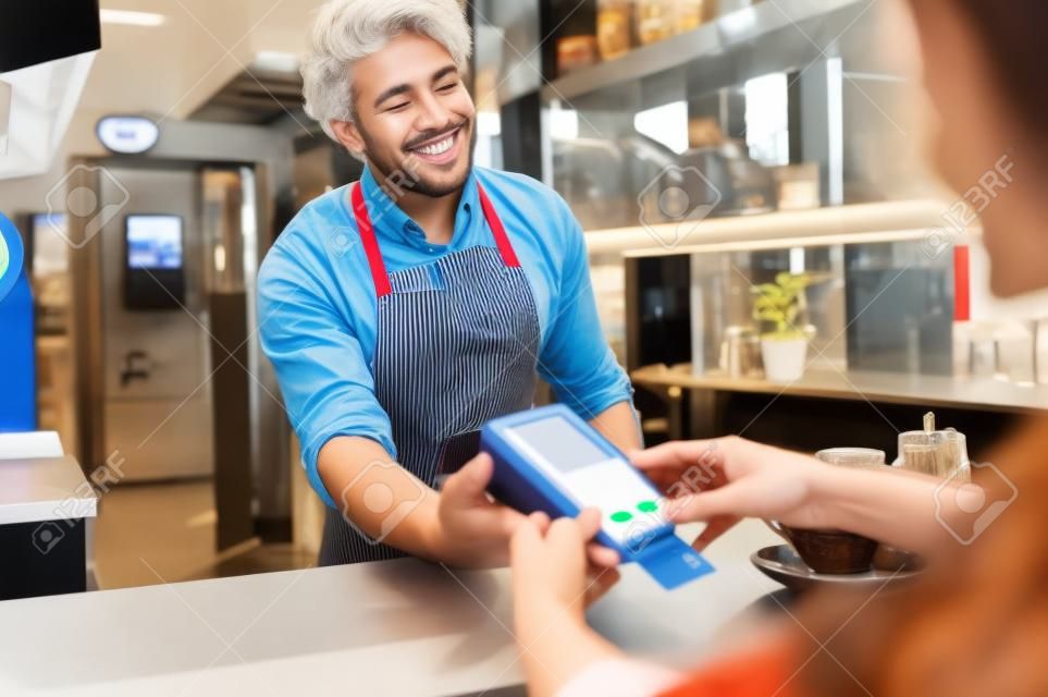 Mujer pagando con tarjeta de crédito e ingresando el código pin en el lector sostenido por un barista sonriente en la cafetería Cliente que utiliza tarjeta de crédito para el pago. Cajero maduro con delantal aceptando pagos a través de tecnología nfc.