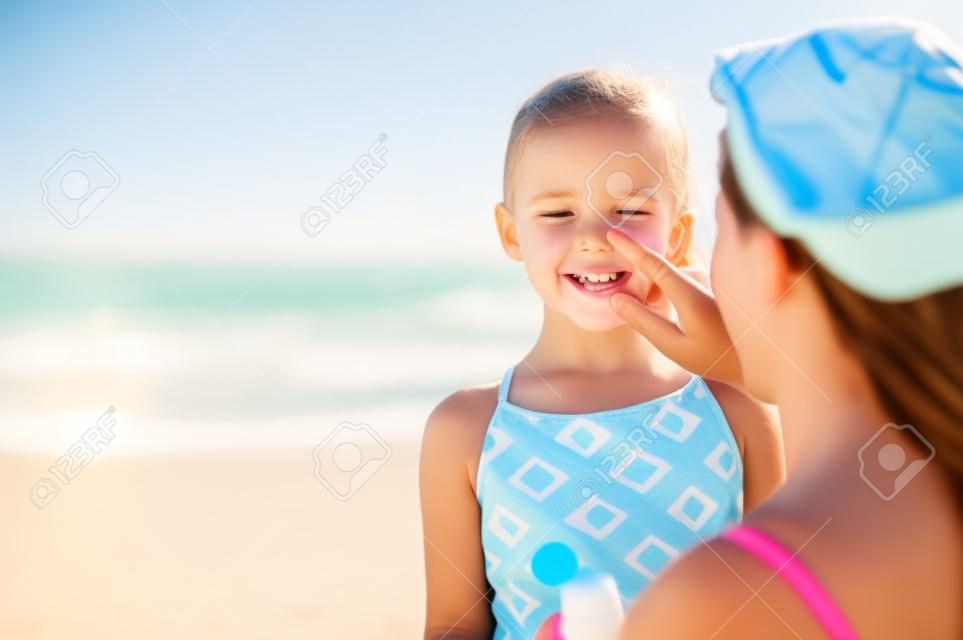 Madre joven que aplica protector solar protector en la nariz de la hija en la playa. Mano de mujer poniendo crema solar en la cara del niño. Niña linda con bloqueador solar en la playa con espacio de copia.