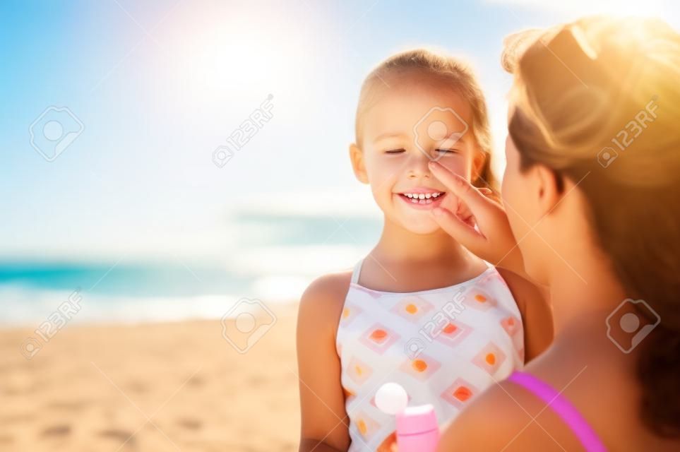 Madre joven que aplica protector solar protector en la nariz de la hija en la playa. Mano de mujer poniendo crema solar en la cara del niño. Niña linda con bloqueador solar en la playa con espacio de copia.