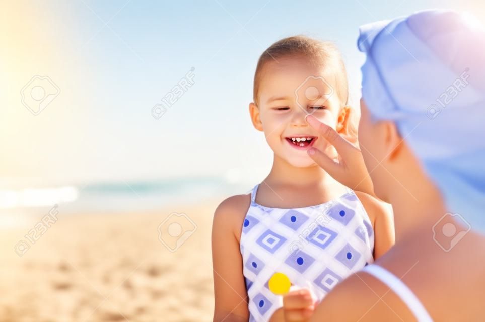 Jovem mãe aplicando protetor solar protetor solar no nariz da filha na praia. Mão da mulher colocando loção solar no rosto da criança. Menina bonita com protetor solar à beira-mar com espaço de cópia.