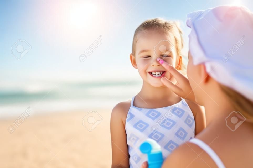 Młoda matka stosowania ochrony przeciwsłonecznej na nosie córki na plaży. Ręka kobiety wprowadzenie balsam do opalania na twarzy dziecka. Cute dziewczynka z kremem przeciwsłonecznym nad morzem z miejsca na kopię.