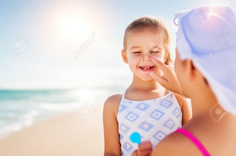 Giovane madre che applica crema solare protettiva sul naso della figlia in spiaggia. Mano della donna che mette la lozione solare sul viso del bambino. Bambina sveglia con crema solare in riva al mare con copia spazio.