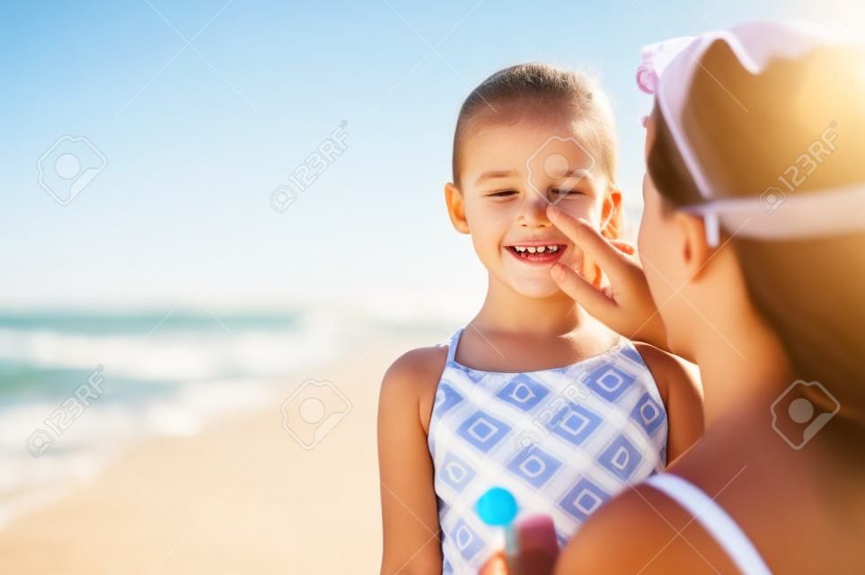 Jonge moeder aanbrengen beschermende zonnebrandcrème op dochter neus op het strand. Vrouw hand zetten zon lotion op kind gezicht. Schattig klein meisje met zonnebrandcrème aan zee met kopieerruimte.