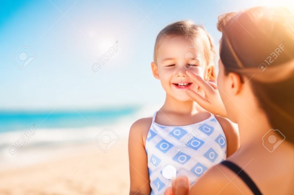 Giovane madre che applica crema solare protettiva sul naso della figlia in spiaggia. Mano della donna che mette la lozione solare sul viso del bambino. Bambina sveglia con crema solare in riva al mare con copia spazio.