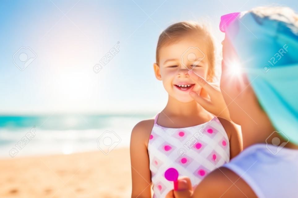 Junge Mutter, die am Strand schützende Sonnencreme auf die Nase der Tochter aufträgt. Frauenhand, die Sonnenlotion auf Kindergesicht setzt. Nettes kleines Mädchen mit Sonnencreme am Meer mit Kopienraum.