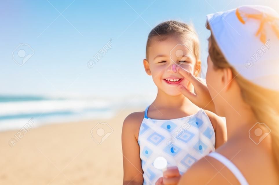 Junge Mutter, die am Strand schützende Sonnencreme auf die Nase der Tochter aufträgt. Frauenhand, die Sonnenlotion auf Kindergesicht setzt. Nettes kleines Mädchen mit Sonnencreme am Meer mit Kopienraum.