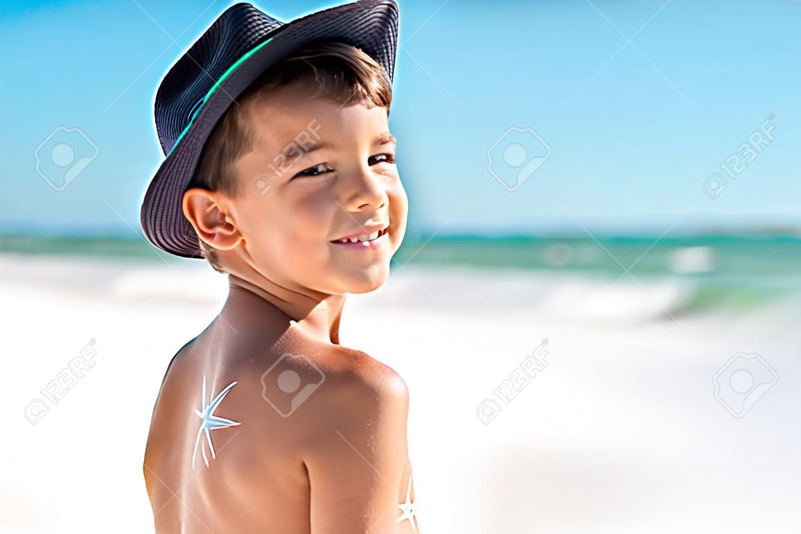 Netter Junge, der am Strand mit Sonne steht, die mit Sonnencreme auf der Schulter gemacht wird und Kamera betrachtet. Glückliches lächelndes Kind mit Feuchtigkeitscreme auf dem Rücken am Strand mit blauem Panamahut. Kleines stolzes Kind, das Urlaub auf See an einem sonnigen Tag mit Kopienraum genießt.