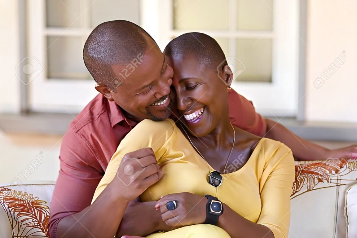 Mature black couple embracing on sofa while looking to each other. Romantic black man embracing woman from behind while laughing together. Happy african wife and husband loving in perfect harmony.