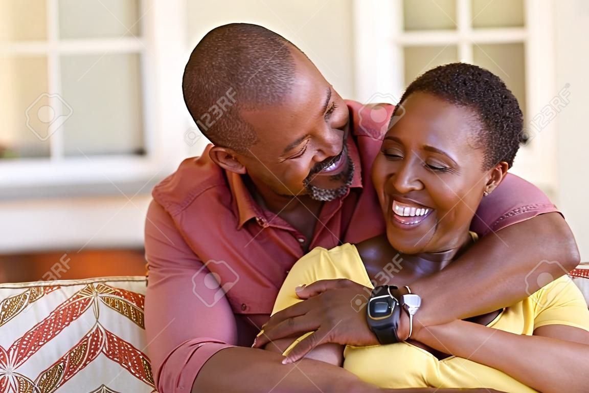 お互いを見ながらソファに抱き合う成熟した黒いカップル。一緒に笑いながら後ろから女性を抱きしめるロマンチックな黒人男性。完璧な調和で愛する幸せなアフリカの妻と夫。