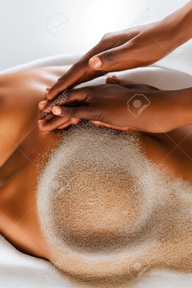 Primer plano de una mujer masajista aplicando exfoliante para pelar a la mujer africana en el spa. Manos del terapeuta aplicando sal de roca de exfoliación en niña en el centro de bienestar para exfoliación corporal. Señora acostada en la mesa de spa para terapia de tratamiento corporal.