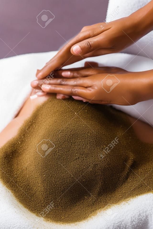 Zbliżenie kobieta masażystka stosowania peelingu do obierania na afrykańskie kobiety z powrotem w spa. Ręce terapeuty, stosując sól kamienną złuszczającą na dziewczynę z powrotem w centrum odnowy biologicznej do peelingu ciała. Pani leżąc na stole spa do terapii ciała.